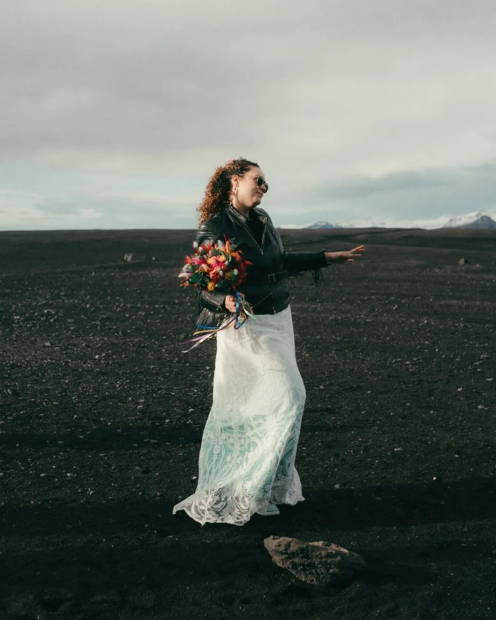 Anna dancing on a black sand beach in Iceland in a lace wedding dress and leather jacket, holding a bright bouquet