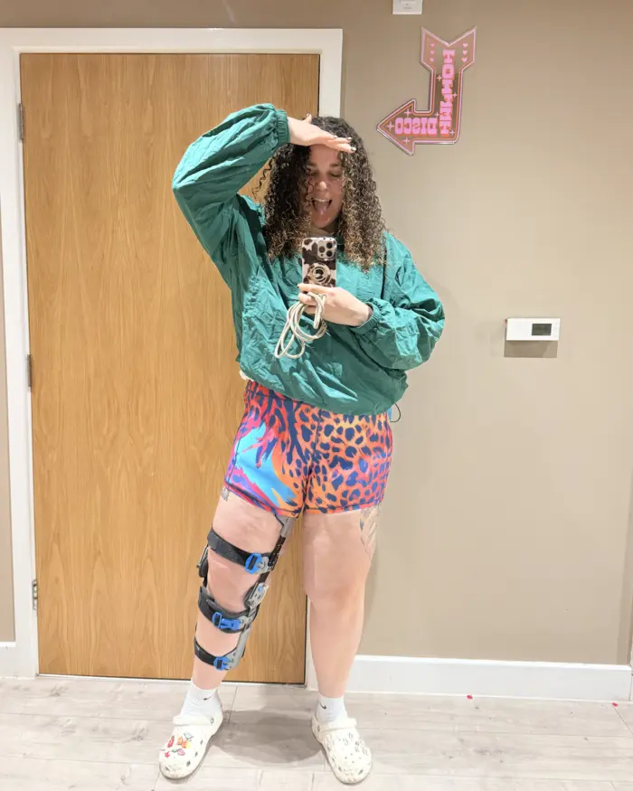 Anna, founder of My Name Changed, pulling a silly face in a mirror selfie, wearing a green jacket, leopard print shorts and a knee brace