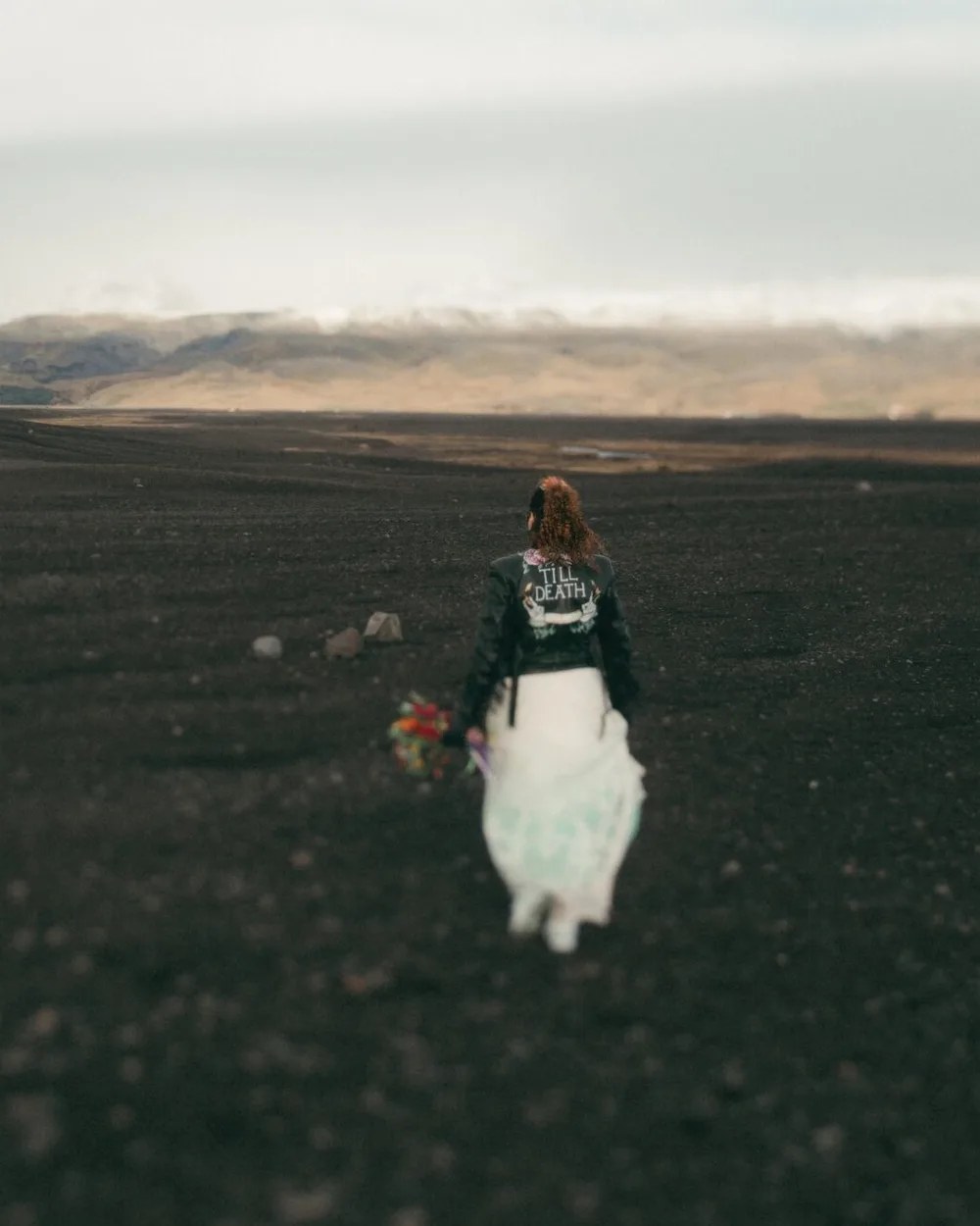 Anna walking away across black sand in Iceland, holding a colourful bouquet