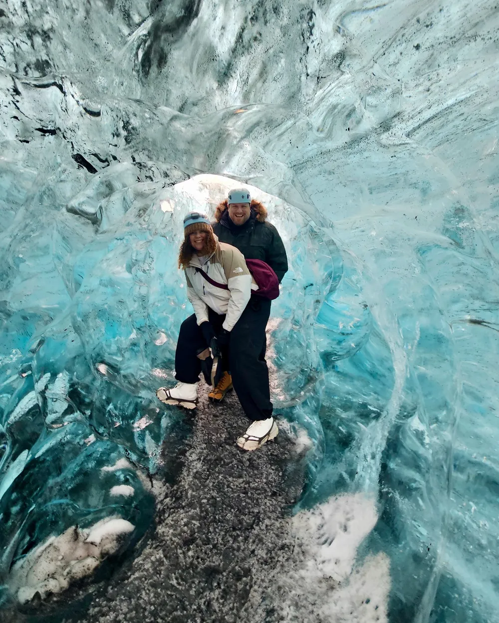 Anna and her partner grinning inside a blue glacier ice cave in Iceland, wearing helmets and crampons