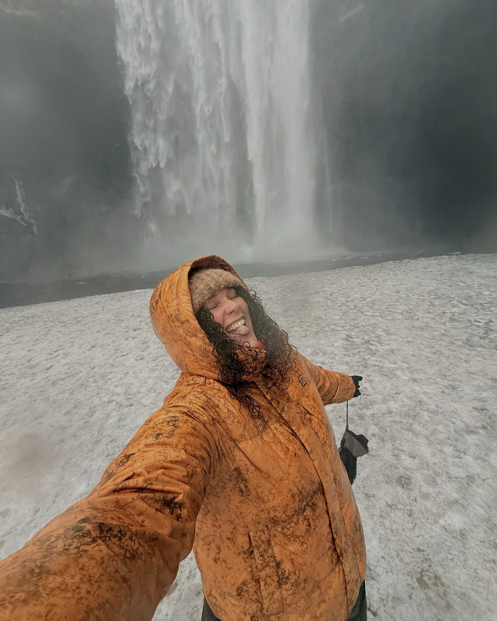 Anna laughing in a mud-splattered orange hooded jacket in front of a huge waterfall in Iceland